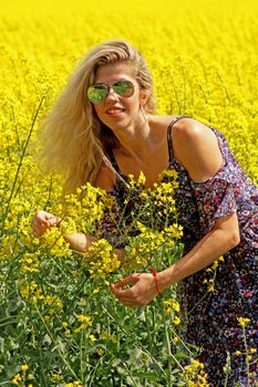 Blonde peasant girl in floral print dress and sunglasses on the yellow blossoming rapeseed field