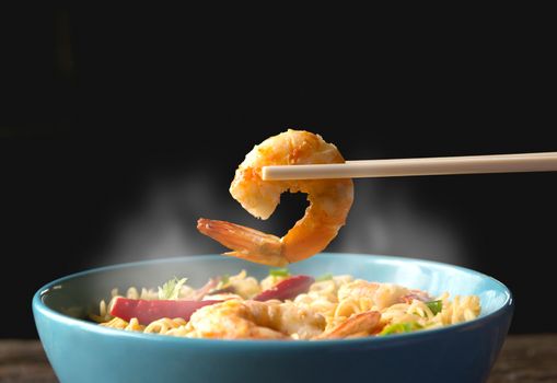 Hand using chopsticks pickup shrimp with instant noodles with smokes isolated on black background.