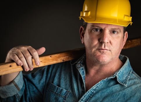 Serious Contractor in Hard Hat Holding Plank of Wood With Dramatic Lighting.