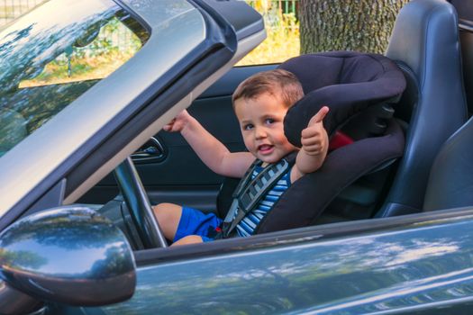 Happy laughing child in a convertible sits in the car child seat and shows the thumbs up.