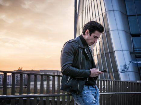 One handsome young man in urban setting in modern city, standing, wearing black leather jacket and jeans