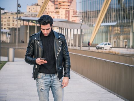 One handsome young man in urban setting in modern city using cellphone, standing and walking, wearing black leather jacket and jeans