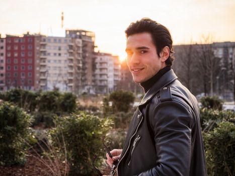 One handsome young man in urban setting in modern city, standing, wearing black leather jacket and jeans, looking at camera, at sunset