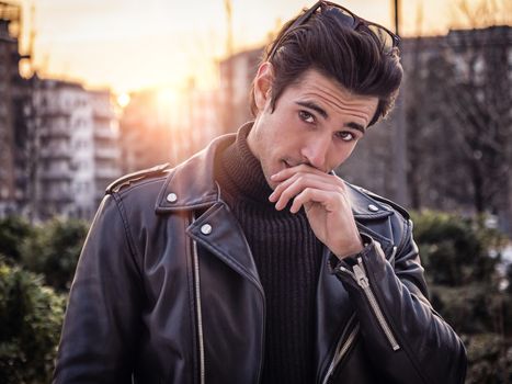 One handsome young man in urban setting in modern city, standing, wearing black leather jacket and jeans, looking at camera, at sunset