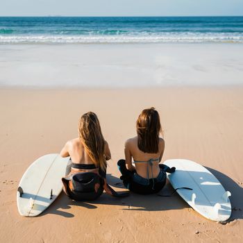 Two beautiful female friends at the beach sitting on the sand with her surfboards while looking to the ocean