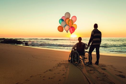 Happy couple at the beach, where woman is on a wheelchair holding balloons on her hands