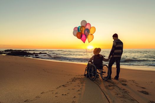 Happy couple at the beach, where woman is on a wheelchair holding balloons on her hands
