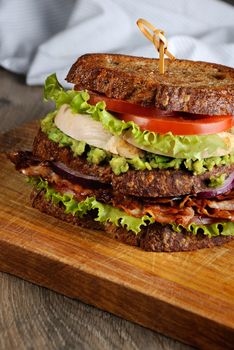 A double sandwich made of Greek bread with slices of fried bacon, avocado, boiled chicken breast, and tomatoes.