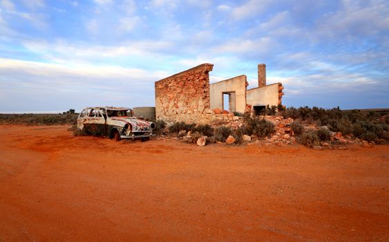 Crumbling old stone home and rusty old car sit on the red ochre earth of remote outback Australia