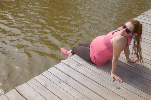 Pregnant woman with pink singlet is sitting on a wooden quay by the water.