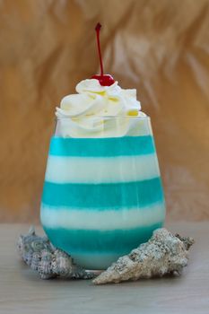 Blue striped jello dessert in glass with whipped cream and red candied cherry on top, seashells, sea vacation concept