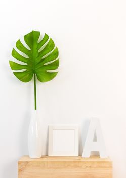 Elegant home decor with white picture frame, green tropical leaf in a vase, and letter A on wooden surface.