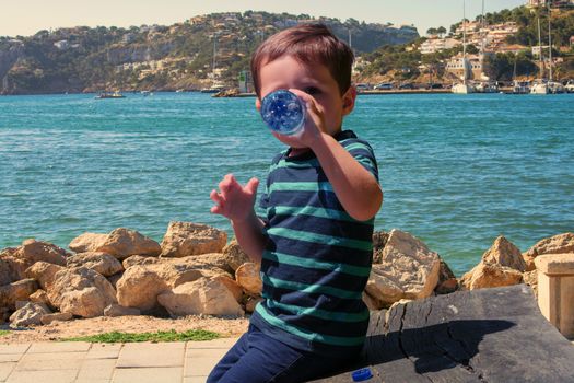 Thirsty little boy sitting on a wooden bench and drinking mineral water. In the background a Mediterranean port.