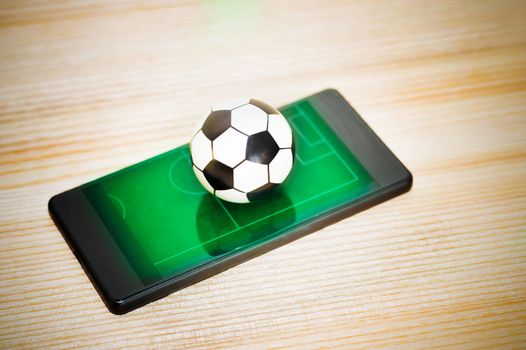 A toy soccer ball on a smartphone with a picture of a green field. Concept of the game of football