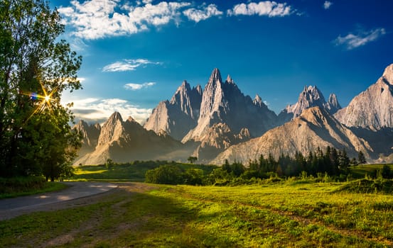 composite landscape with rocky peaks at sunset. beautiful mountainous scenery with road going through grassy hills in to the distance. sun shine through trees