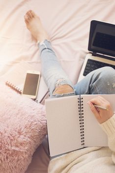 Lifestyle soft image of pretty young woman sitting in her cozy bed and making notes to her diary. Feminine workplace concept. Blogger working.