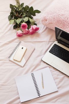 Roses, laptop, notebooks and smartphone in bed on pink sheets. Freelance fashion home femininity workspace in flat lay style.