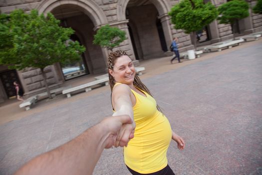 Laughing pregnant woman holding male hand is taking a walk outdoors.