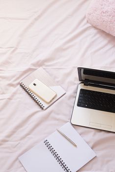 Laptop, notebooks and smartphone in bed on pink sheets. Freelance fashion home femininity workspace in flat lay style.