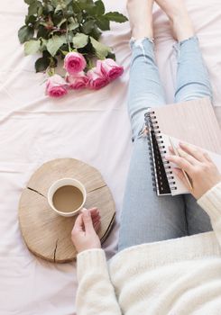 Young Caucasian woman in bed early in the morning, wearing distressed jeans and a white sweater and holds a cup of coffee. Roses and notebooks around. Cozy blogger mornings