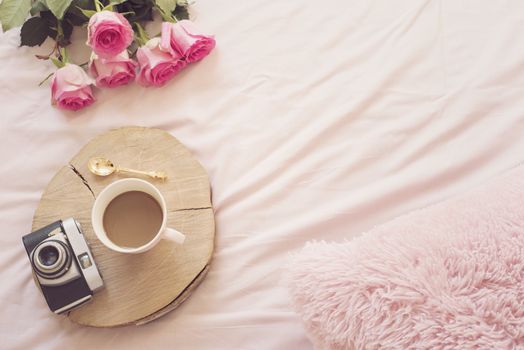 Coffee, old vintage camera in bed on pink sheets. Roses and notebooks around. Freelance fashion home femininity workspace in flat lay style.