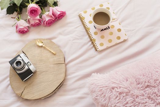 Coffee, old vintage camera in bed on pink sheets. Roses and notebooks around. Freelance fashion home femininity workspace in flat lay style