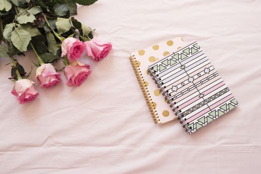 Floral frame with stunning pink roses and notebooks on pink bed sheets in the bedroom. Freelance workspace. Wedding, gift card, valentine's day or mother's day background