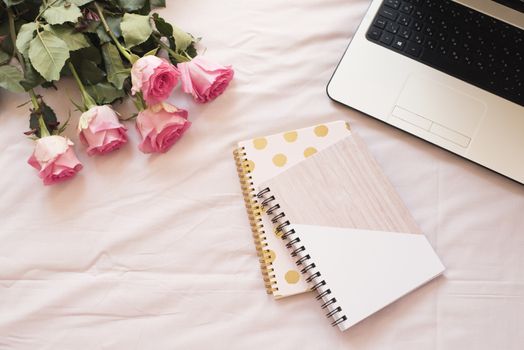 Notebooks, laptop, bouquet of roses in bed on pink sheets. Freelance fashion home femininity workspace in flat lay style.