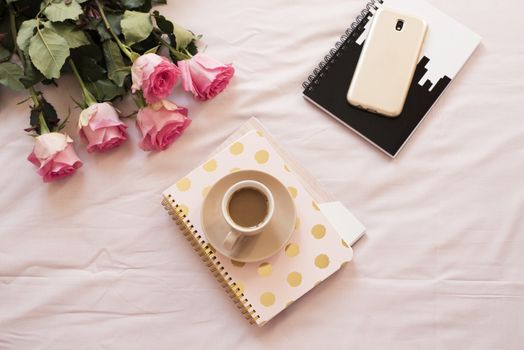 Coffee in bed on pink sheets. Roses, notebooks, gold smartphone around. Freelance fashion home femininity workspace.