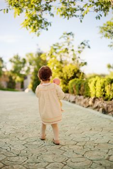 Toddler girl in the back, dressed in a bright dress with a wreath of flowers on his head and holding a doll. Little girl walking down an stone alley in a garden with flowers and trees.