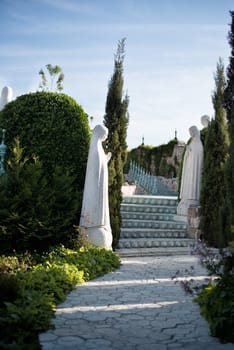 Stone walkway.  Old statues of wife, women, Mary surrounds. Alley in beautiful garden with flowers and trees around. Summer in the garden