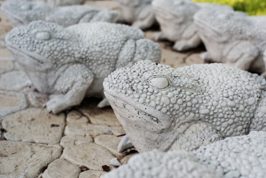 Limestone statues of frogs. Stone walkway. Frogs on alley in beautiful garden with flowers and trees around. Summer in the garden