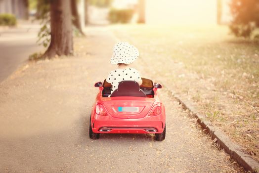  Little girl dressed in white blackheads shirt and hat, driving a red toy car near the park. Baby girl, child  playing with car. Sun, sun haze, glare. 