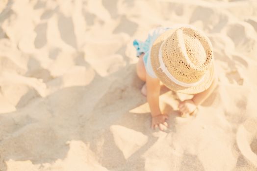 Baby girl child with straw hat and blue dress playing with sand at the beach in summer. Little girl sitting on the shore of the sea. Sun, sun haze, glare