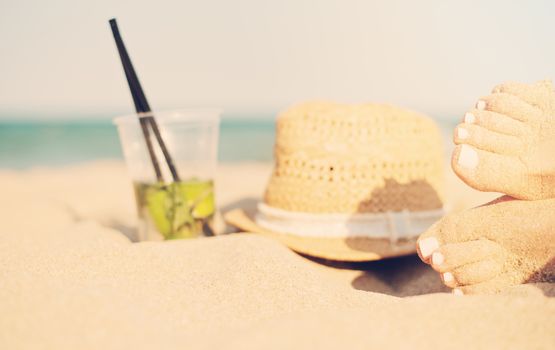 Leisure in summer - Beautiful of sexy women feet, female legs on sandy beach with hat and mojito cocktail. Mojito cocktail on the beach and straw hat. Sun, sun haze, glare