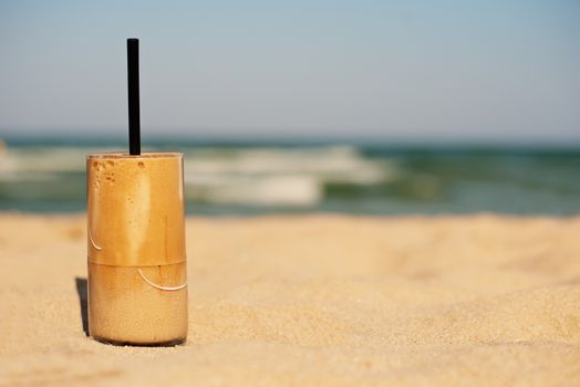 Frappe, ice coffee on the beach. Summer iced coffee (frappuccino, frappe or latte) in a tall glass on sand background - beach bar holiday cold beverages menu. Layout with free text space. Copy space on right