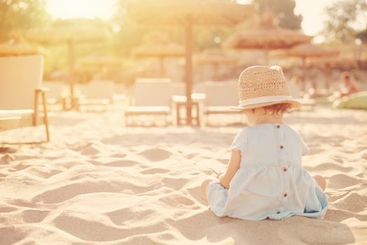 Baby girl child with straw hat and blue dress looking at the phone on a sunny day. Little girl sitting back on the shore of the sea. Sun, sun haze, glare