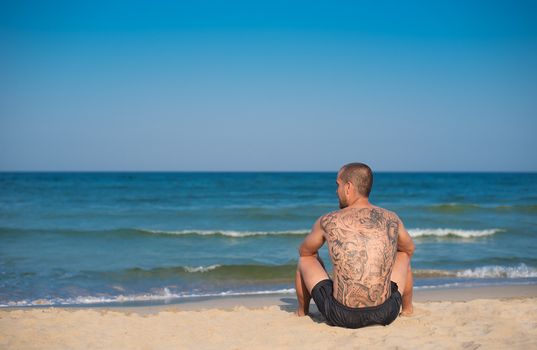  A young man with a large tattoo on his back sitting on the beach overlooking the sea. Copy space on left 