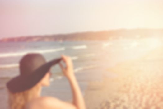 Blurred picture of beautiful young lady in black hat on the beach. Pretty girl looking at distance on blurred lens flare beach, sea background. Sun, sun haze, glare. Copy space on right