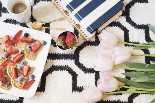 Belgian waffles on the floor. Coffee, berries, notebooks on Scandinavian rug. Pink Tulips and Gold Spoons. White black pattern and gold theme. Lifestyle concept