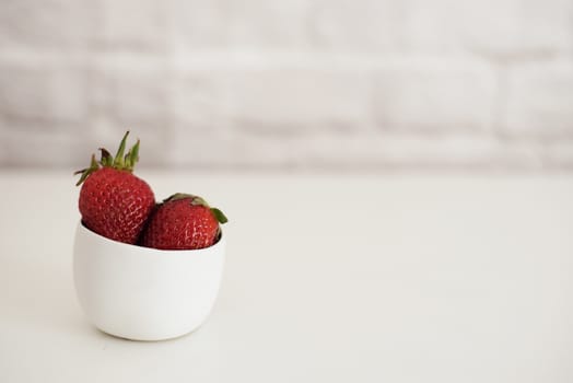Fresh strawberries in a small white bowl. White background, brick wall. Bright composition