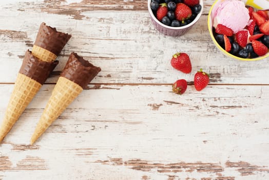 Pink Ice Cream served with berries - strawberries and blueberries in a yellow bowl. Waffle cones with chocolate. Light Rustic Wooden Background