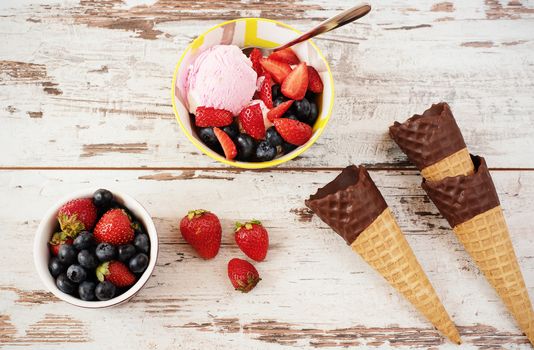 Pink Ice Cream served with berries - strawberries and blueberries in a yellow bowl. Waffle cones with chocolate. Light Rustic Wooden Background