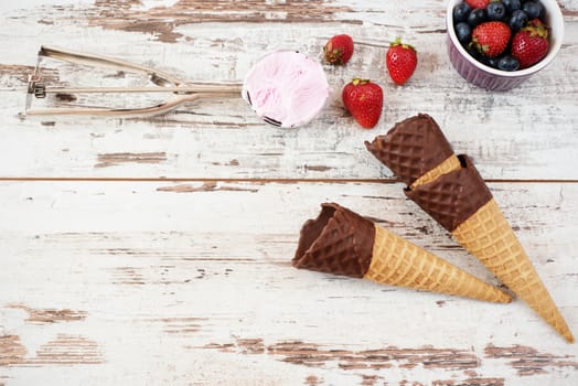 Pink Ice Cream served with berries - strawberries and blueberries in a yellow bowl. Waffle cones with chocolate. Light Rustic Wooden Background