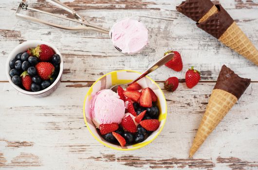 Pink Ice Cream served with berries - strawberries and blueberries in a yellow bowl. Waffle cones with chocolate. Light Rustic Wooden Background