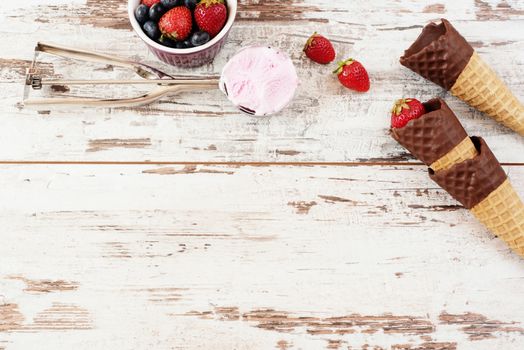 Pink Ice Cream served with berries - strawberries and blueberries in a yellow bowl. Waffle cones with chocolate. Light Rustic Wooden Background