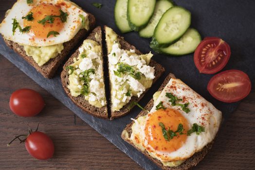 Avocado Toast. Healthy Breakfast. Top View. Homemade Sandwich With Avocado And Fried Eggs, Cherry Tomato And Cucumbers On A Wooden Background. Dark Food Photography