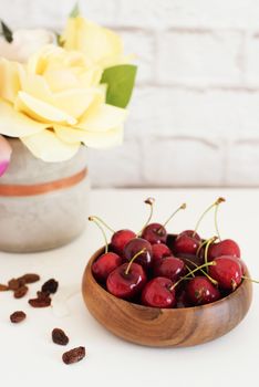 Fresh cherries in a wooden bamboo bowl. Bright Brick Wall Background. Yellow Roses in a Vase. Healthy Lifestyle Concept. Copy Space