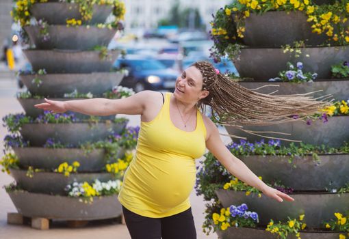 Happy pregnant woman is spinning outdoors in front of big flower pots, hands stretched.