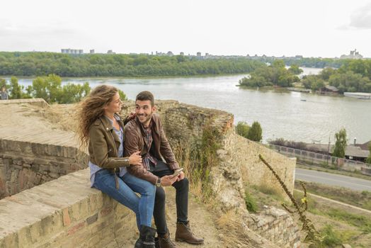 Attractive long haired woman and her boyfriend sitting on the wall and using their smart phone
